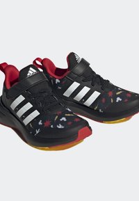 Scarpe da ginnastica nere con una tomaia decorata, caratterizzata da accenti rossi e gialli, tre strisce bianche e una suola in gomma nera con una base gialla.