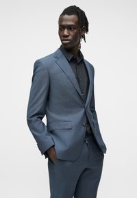 Homme aux dreadlocks portant un costume bleu et une chemise noire, debout les mains dans les poches, regardant sur le côté sur un fond uni.