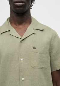 Άνδρας που φοράει ανοιχτό πράσινο πουκάμισο με κοντά μανίκια, καταπράσινο γιακά τύπου καμπ, και μικρό ορθογώνιο λογότυπο κόκκινο, λευκό και μπλε στην τσέπη.