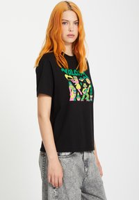 Zwart T-shirt met korte mouwen met kleurrijk grafisch ontwerp en de tekst "WOLC." Het model draagt het met lichtgrijze jeans, wat een ontspannen pasvorm laat zien.