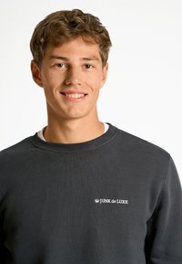 Sudadera gris oscuro con textura acanalada y cuello redondo. Presenta un pequeño logo blanco "JUNK de LUXE" en la parte izquierda del pecho.