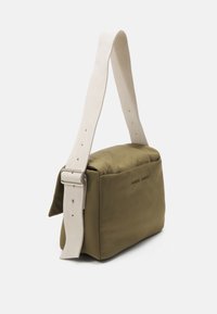 Bolso rectangular verde oliva con una correa gruesa color crema, con una textura suave y un cierre metálico. Logo grabado en la parte frontal.