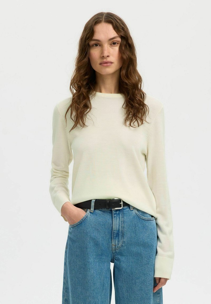 Femme aux longs cheveux ondulés portant un haut crème à manches longues rentré dans un jean bleu taille haute avec une ceinture noire, debout avec une main dans la poche.