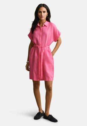 Vrouw staand met handen in de zakken, gekleed in een kortarmige roze jurk met ceintuur en zwarte veterschoenen, tegen een effen achtergrond.