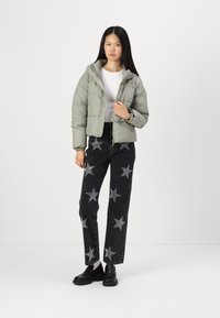 Doudoune vert clair avec capuche et fermeture éclair. Portée avec un jean large noir orné de motifs d'étoiles argentées. Des chaussures noires complètent la tenue.