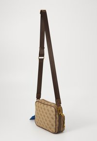 Geantă crossbody bej cu un model de logo maro, curea ajustabilă maro și fermoare cu ton de aur, stand bine vertical pe un fundal alb.