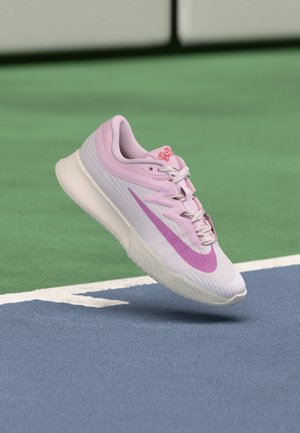 Chaussure de course Nike rose et blanche avec un swoosh violet flottant au-dessus d'une surface de terrain de tennis bleue et verte.