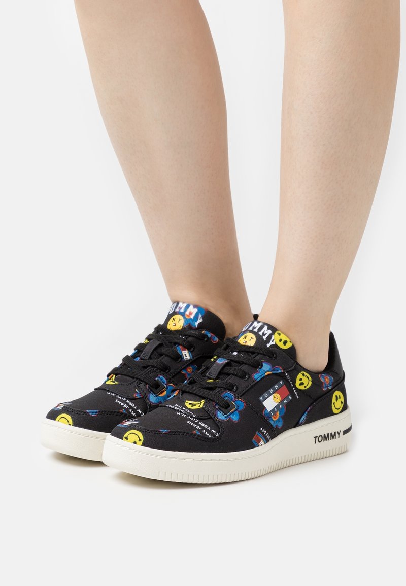 Zapatillas negras de caña baja con caras sonrientes amarillas, flores azules y logotipos blancos de "Tommy Jeans", usadas sin calcetines sobre un fondo blanco.