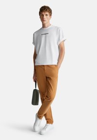 Mladý muž v bílem tričku s nápisem "NUMBERS", hnědých kalhotách, bílých teniskách, držící šedou tašku, stojící s překříženýma nohama na bílém pozadí.