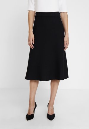 Femme portant une jupe noire mi-longue en ligne A, un haut blanc à manches coudées et des talons noirs à bout pointu, debout sur un fond blanc uni.