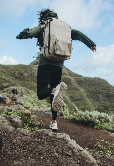 Zaino beige con più scomparti, indossato da una persona con una giacca verde e leggings neri, che corre su un sentiero roccioso circondato da vegetazione.