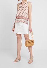 Blusa senza maniche rosa con motivo paisley rosso ripetuto, abbinata a una mini gonna in denim bianco e a una borsa in legno a righe. Sandali con tacco blocco color beige.