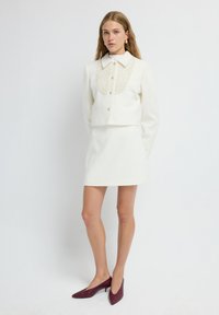 White, textured two-piece outfit featuring a cropped jacket with a large collar and buttons, paired with a matching mini skirt and burgundy heels.