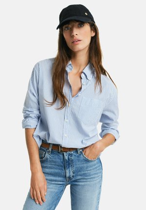 Light blue and white striped button-up shirt, rolled sleeves, paired with light blue jeans and a black cap with a gold accent.