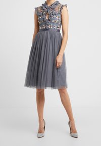 Robe florale grise avec un col transparent à volants, manches courtes et jupe en tulle à pois. Portée avec des chaussures à talons hauts argentées.