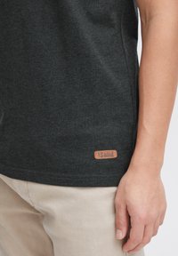 Camiseta de color gris oscuro hecha de una tela suave con una textura lisa, que presenta un parche de logo de cuero marrón en el dobladillo y un diseño ajustado.