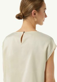 Jasno beżowa bluzka z tylnym wycięciem w kształcie łezki i zapięciem na pojedynczy guzik. Miękka, gładka tkanina, krótkie rękawki i luźny krój.