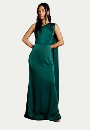 Donna con lunghi capelli scuri che indossa un abito lungo senza maniche in raso verde smeraldo con dettaglio mantello, in piedi con le mani sui fianchi.