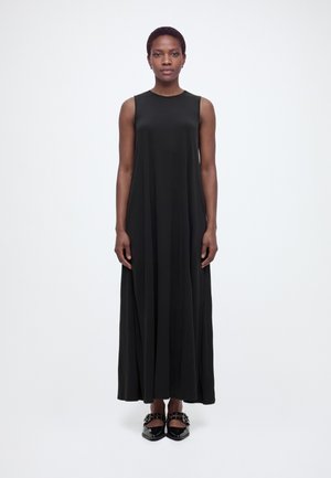 Frau steht aufrecht und trägt ein ärmelloses schwarzes Maxikleid und schwarze spitze Schuhe mit dekorativen Riemen vor weißem Hintergrund.