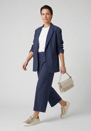 Kobieta idzie, ubrana w granatowy garnitur z podwiniętymi rękawami, białą koszulę, złote trampki i niesie beżową torebkę.