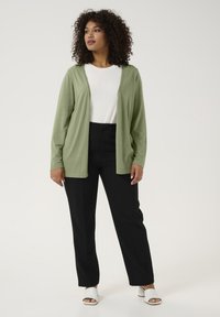 Cardigan verde deschis, bluză albă și pantaloni negri. Cardiganul are mâneci lungi și un front deschis, completat de sandale albe cu toc.