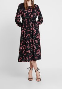 Black floral dress with pink flowers, long sleeves, elastic waist, and a midi length. Paired with strappy black high-heeled sandals.