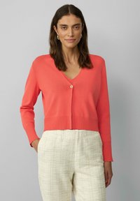 Korallenfarbener Cardigan mit V-Ausschnitt, einfacher Knopföffnung und geripptem Saum. Trägt man über einer hellen, strukturierten Hose, mit langen Ärmeln und weichem Strickmaterial.