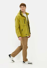 Gul vandtæt jakke med flere lommer og sorte lynlåse, parret med beige bukser og hvide sneakers med sorte detaljer.