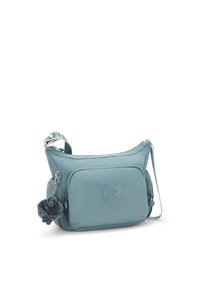 Teal-krossbody laukku, joka on valmistettu pehmeästä kankaasta. Laukussa on etutasku, pyöreä logo ja vetoketjuun kiinnitetty pörröinen pom-pom-koriste.