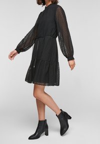 Black sheer dress with textured polka dots, long sleeves, and a tiered hem. Paired with black heeled ankle boots.