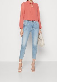 Korallinvärinen blus med långa ärmar och knytning vid halsen, tillsammans med ljusblå skinny jeans. Accessoarer inkluderar en krämfärgad handväska och vita klackar.