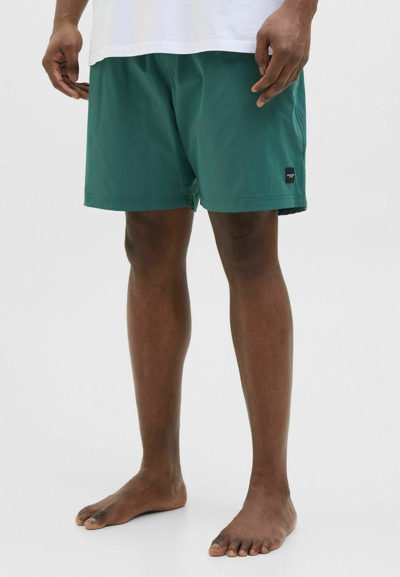 Shorts de bain verts en tissu léger, longueur mi-cuisse, avec une petite étiquette de marque noire sur le côté et une taille élastique.