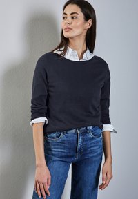 Vrouw in een donker trui, wit overhemd met kraag en blauwe spijkerbroek staat tegen een lichtgrijze muur, kijkt naar rechts met ontspannen armen.