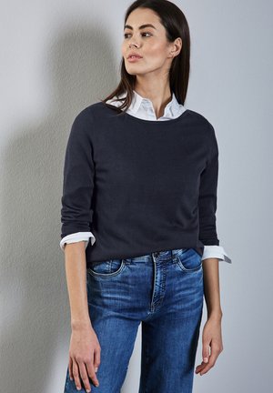 Maglione blu scuro con scollatura rotonda, indossato sopra una camicia bianca con colletto. Abbinato a jeans in denim blu chiaro. Tessuto dalla consistenza liscia.