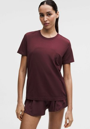 Naine, kellel on tume juuksed tagant seotud, kannab bordoopunast lühikeste varrukatega T-särki ja sobivaid lühikesi pükse, seisab heleda taustaga vastu.