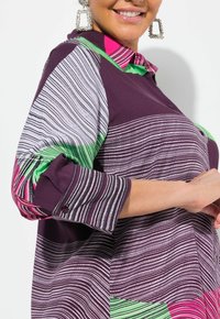 Camicia multicolore con strisce viola, verdi e rosa, colletto con bottoni e maniche arrotolate. Tessuto morbido con motivi distintivi.