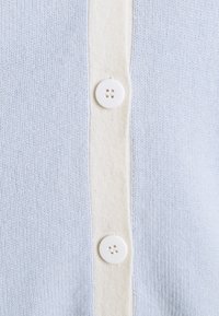 Ljusblå, stickad kofta med en krämfärgad knappslå och vita knappar med fyra hål. Mjuk textur och enkel design.