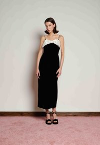 Μαύρο βελούδινο φόρεμα με λευκή λεπτομέρεια σατέν φιόγκου στο μπούστο, λεπτές τιράντες και μήκος μέχρι το μέσο της γάμπας. Φορεμένο με μαύρες πέδιλες με λουριά.