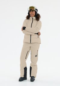 Beige Skijacke und -hose mit schwarzen Akzenten. Die Jacke hat einen hohen Kragen und Reißverschlusstaschen. Das Modell trägt einen Helm und eine Skibrille.