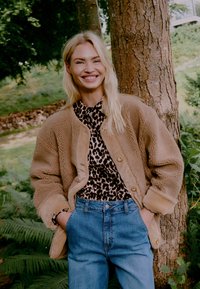 Tan fleecetop met grote knopen over een zwart en bruin panterprintshirt, gecombineerd met lichtblauwe high-waisted jeans.