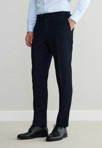 Navy pinstriped trousers made from a woven fabric, featuring side pockets and a button closure, paired with black lace-up shoes.