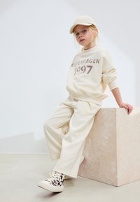 Sweat-shirt et pantalon de survêtement de couleur crème avec l'inscription "COPENHAGEN 1997", assortis à une casquette beige et des baskets blanches à motif.