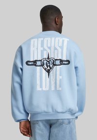 Gaiši zils džemperis ar apaļu kakla izgriezumu, ar lieliem melniem grafiskiem uzrakstiem "PRETOT MĪLESTĪBA" un sirds dizainu aizmugurē.