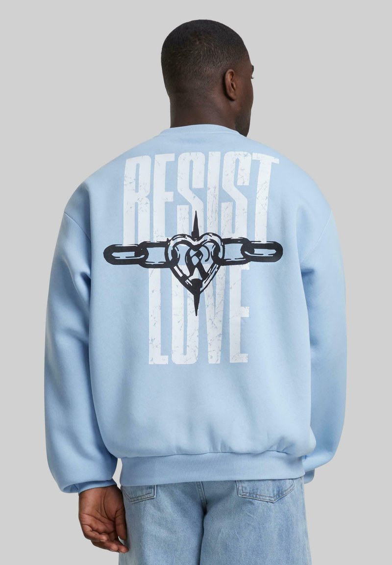 Gaiši zils džemperis ar apaļu kakla izgriezumu, ar lieliem melniem grafiskiem uzrakstiem "PRETOT MĪLESTĪBA" un sirds dizainu aizmugurē.