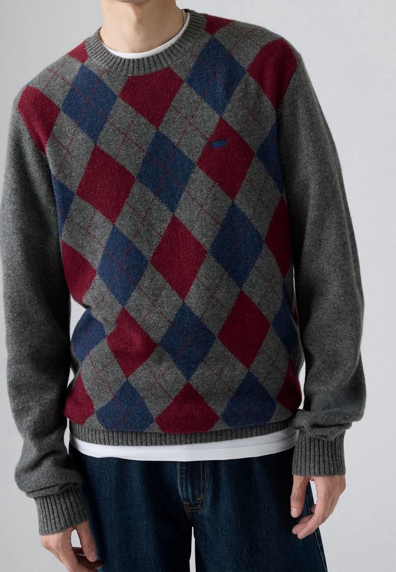 Pull en laine grise arborant un motif losangé rouge et bleu, avec un col et des poignets côtelés, porté sur une chemise blanche.