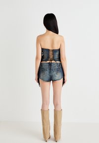 En person står med ryggen til kameraet, iført en stroppeløs denimtop med snøring og høyhælede beige støvler. Shorts er synlige.