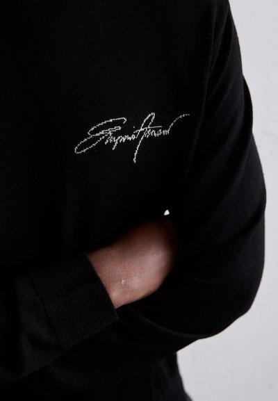 Camisa negra de manga larga con un bordado blanco del logo en el pecho. La tela parece suave, con puños y dobladillo acanalados.