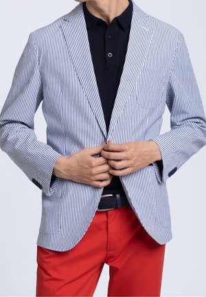Blazer rayé en blanc et marine, avec une coupe ajustée, une poche appliquée et un col cranté, associé à un pantalon rouge et un polo noir.