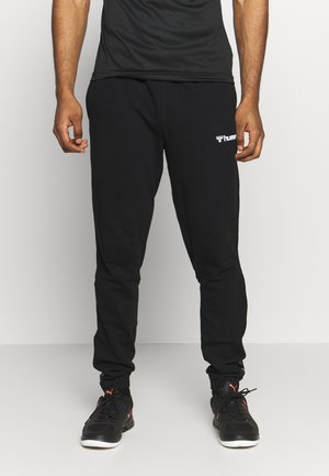 Pantalons de jogging noirs en coton avec des poignets élastiques et un petit logo blanc sur la cuisse droite. Portés avec des chaussures de sport noires.