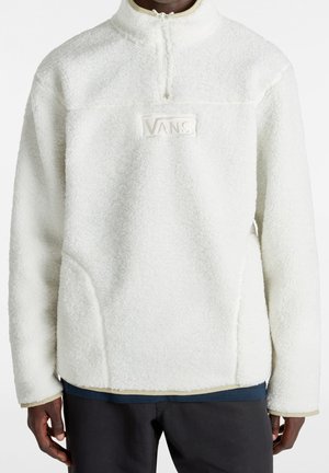 Osoba ubrana w biały, teksturowany polar z suwakiem do kwadratu i naszywką z logo Vans na piersi.